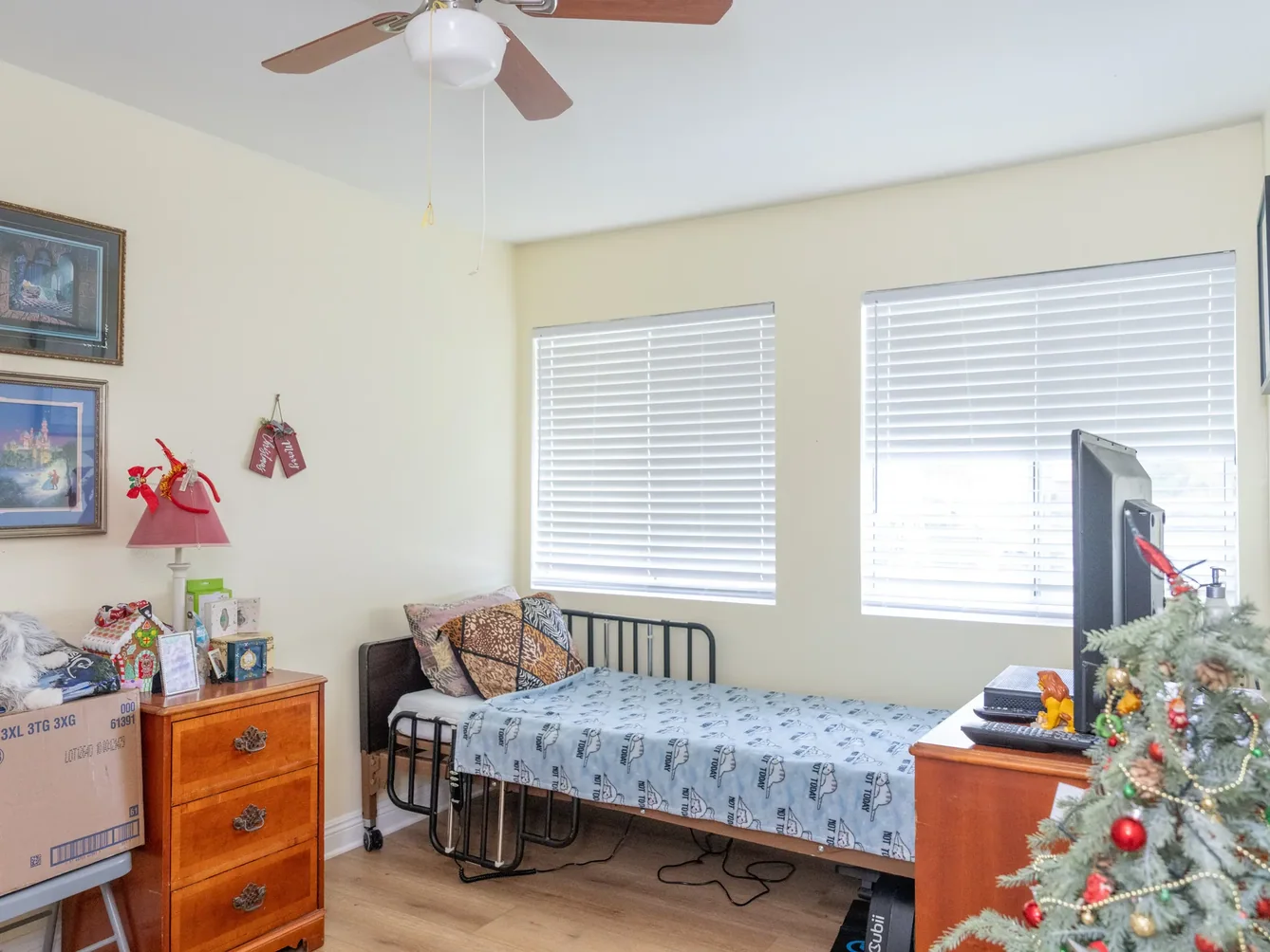 Bright accessible bedroom featuring adjustable hospital bed, wooden dresser, dual windows with blinds, and ceiling fan in residential care facility